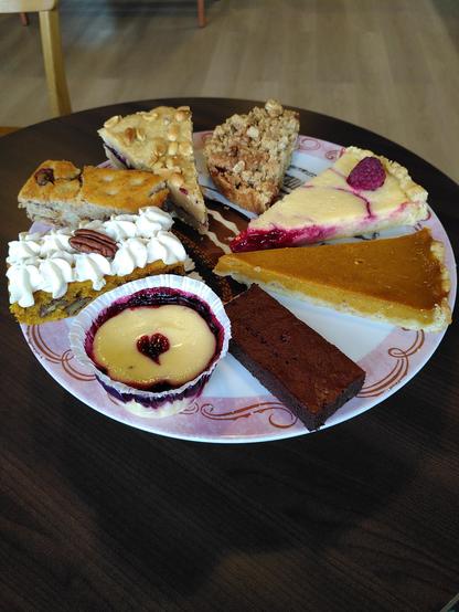 Auf einer rosafarbenen, runden befinden sich acht verschiedene Leckereien, u. a. ein Frischkäsetörtchen mit eingebackenem Holunderbeer-Apfel-Püree, ein klassischer Schokobrownie, ein Stück cremig-würziger Kürbiskuchen und eine Zitronentarte mit Himbeerpüree.