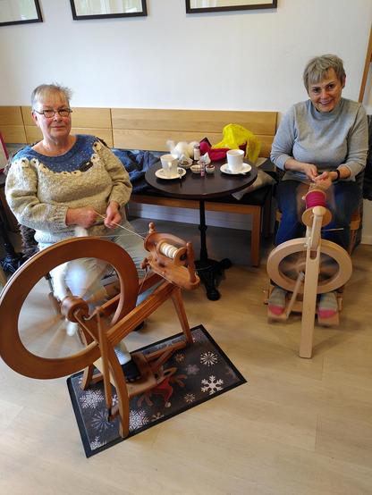 Vor einer Sitzecke im Café sitzen zwei Frauen mit kurzen, grauen Haaren auf je einem Stuhl. Vor ihnen befinden sich zwei Spinnräder aus Holz. 
Auf dem Tisch dahinter steht etwas Geschirr, u. a. zwei Becher mit Kräutertee.