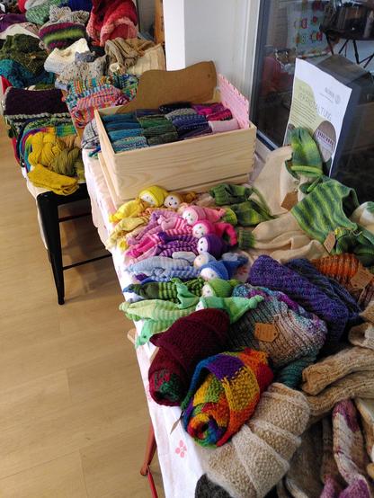 Blick auf viele verschiedene, in bunten Farben gestrickte und gehäkelte Schals, Socken und Püppchen. Diese sind in den Farben des Regenbogens auf dem Tisch arrangiert.