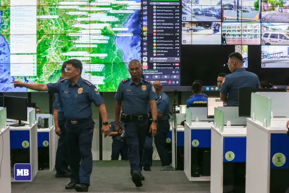 Two high-ranking Philippine National Police (PNP) officers walk through a high-tech command center in uniform. In the background, a massive monitoring wall is dominated by the communication software Discord (and a map), displaying active voice chats in the server. The voice chats are populated with accounts with their webcam turned on, streaming ACTUAL CCTV FOOTAGE. Dispatchers sit at white cubicles facing the screens.