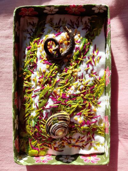 As mall rectangular open box, embellished with a floral fabric is used as a frame for a wild, impressionistic embroidery in nuances of green, red, and yellow. It's like a plant structure. At the bottom I glued a real empty shell of a snail, at the top a spiraling wooden part of vine.