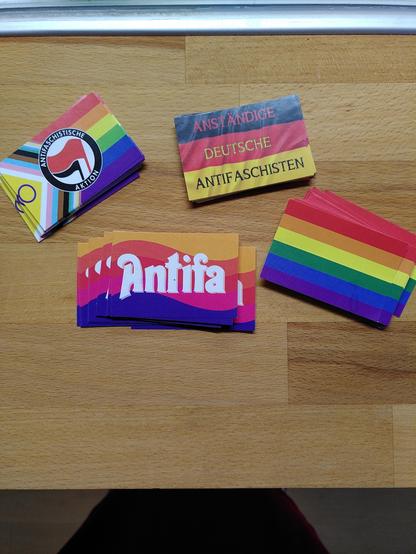 Auf einem hellbraunen Holztisch liegen vier kleine Stapel mit bunten Aufklebern. Alle sind rechteckig und etwa vier mal zwei Zentimeter groß. Ein Aufkleber trägt die Farben des Regenbogens, ein zweiter den Schriftzug *Antifa* , ein dritter den Schriftzug *Anständige Deutsche Antifaschisten* (dieser prangt auf der Deutschlandfahne) und ein vierter bewirbt die antifaschistische Fraktion auf der LGBTQI+-Fahne.