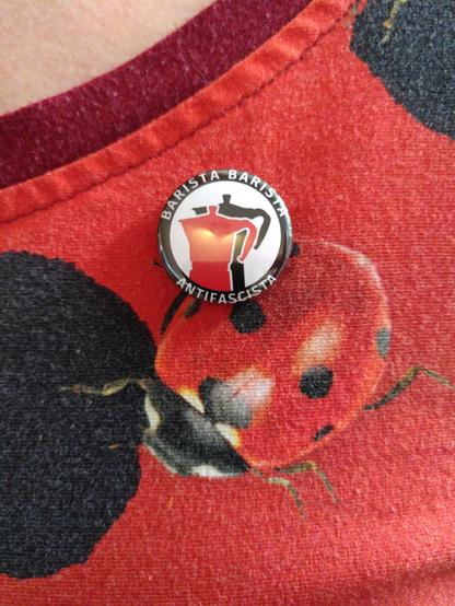 Auf einem Kleid aus rot-schwarz gemustertem Stoff mit einem Marienkäfer darauf prangt eine runde Anstecknadel, die in der Mitte zwei Espressokannen zeigt, eine rote und eine schwarze. Eingerahmt werden diese durch den Schriftzug *Barista, Barista, Antifascista*.