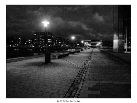 Schwarz-Weiß Nachtaufnahme. Gepflasterter Weg, auf der linke Seite eine Reihe von Straßenlaternen, dramatische Wolken.
