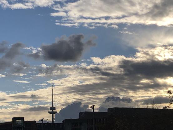 Panorama Ansicht mit Fernsehturm von erhöhtem Standpunkt. Im Gegenlicht sind Gebäudeblöcke am unteren Bildrand nur schwarze Silhouetten. Am Himmel ziehen sich von rechts helle und dunkle Wolken zusammen während links blauer Himmel ist. Sonne hinter den Wolken.
