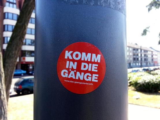 Gemini Beschreibung:
Das Bild zeigt eine Nahaufnahme eines kreisrunden Aufklebers an einem grauen, metallischen Masten im öffentlichen Raum.   Hauptmotiv & AufkleberAufkleber: Auf dem Masten ist zentral ein kreisrunder, roter Sticker angebracht.   Text & Schrift:In dreizeilig gesetzten, weißen Blockbuchstaben steht mittig der Slogan „KOMM IN DIE GÄNGE“.   Darunter ist am unteren Rand in kleinerer weißer Schrift die Internetadresse www.das-gaengeviertel.info zu lesen (eine Initiative zur Erhaltung des historischen Gängeviertels in Hamburg).   Hintergrund & UmgebungMast: Der dunkelgrau/anthrazit beschichtete Metallmast (vermutlich ein Laternen- oder Schildermast) dominiert vertikal die Bildmitte.   Hintergrund: Der unscharfe Hintergrund zeigt eine städtische Straßenszenerie im Tageslicht:Links ist ein mehrstöckiges Wohn- und Geschäftshaus mit dunklen Fensterfronten und einem davor stehenden Baumstamm zu erkennen.   Weiter unten parken Autos am Straßenrand.   Rechts schließt sich eine Bepflanzung/Grünanlage sowie die Silhouette weiterer Wohngebäude an.