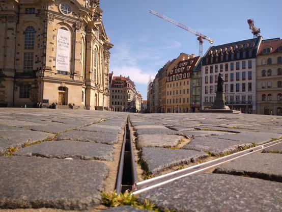 Zwischen den Kopfsteinpflastern sind Sickerrinnen eingelassen, um Regenwasser abzutransportieren. Frosch-Perspektive, aus unmittelbarer Bodenhöhe, so dass in der Flucht eine solche Rinne von der Betrachter:in wegführt. Links steht die Frauenkirche, an der in einem Fenster ein großes Banner hängt, mit der Aufschrift: Selig sind, die Frieden stiften. / Blessed are the peace-makers.