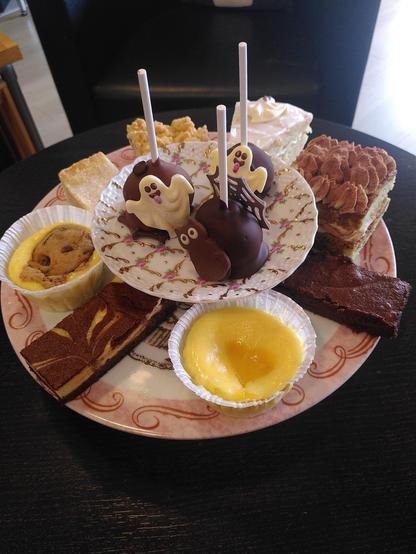 Auf einem runden, dunkelbraunen Holztisch steht eine große, ebenfalls runde Kuchenplatte mit diversen verschiedenen Kuchen und Törtchen darauf, u. a. ein Stück Tiramisútorte, ein Cheesecake mit Lemon Curd (eine Zitronencrème) und ein Brownie mit Frischkäsecrème. In der Mitte thront eine kleine Kuchenplatte mit Fuß, auf der sich drei in dunkle Schokolade getauchten Kuchen am Stiel befinden. Zwei dieser Küchlein sind mit je einem Gespenst aus weißer Schokolade verziert, das dritte mit einer Katze aus dunkler Schokolade.