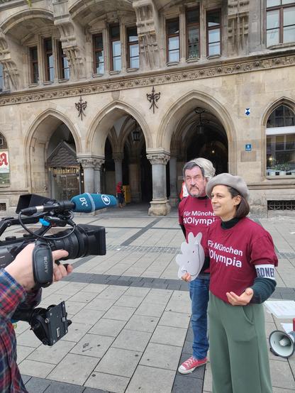 Junge Frau spricht in BR Mikro und Kamera. Dahinter steht Typ mit Södermaske. Beide tragen lila nolympia T-Shirt. Im Hintergrund der Rathauseingang.