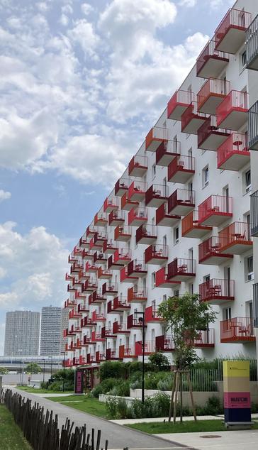 Ein moderner Wohnblock mit einer Fassade aus weißen Wänden und sehr vielen einzelnen Balkonen mit Balkongittern in unterschiedlichen Rottönen. 
Auf fast allen Balkonen stehen ein runder Tisch und ein bis zwei Stühle, diese sind ebenfalls rot.
Die Szene umfasst einen Landschaftsbereich mit Grün im Vordergrund und einem bewölkten Himmel im Hintergrund.