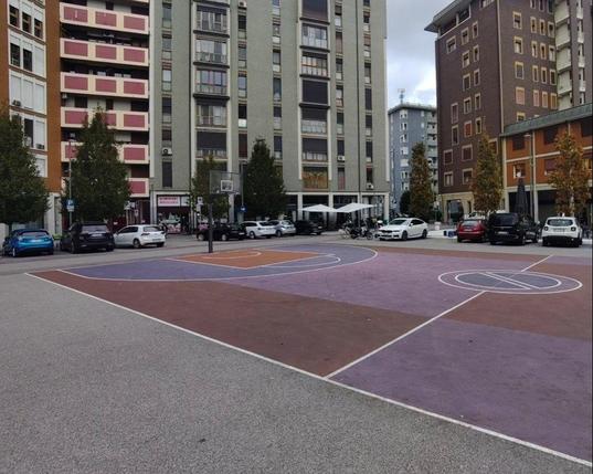 Foto del campo da basket. Il colore del terreno è bordeaux-magenta. Il cerchio centrale del campo è un grosso segnale do divieto di sosta. Intorno al campo sono parcheggiate delle auto. Il piazzale è circondato da palazzi.