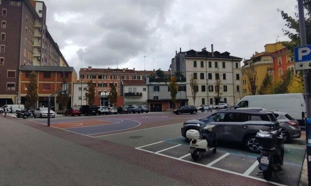 Altra foto del campo da basket. Il colore del terreno è bordeaux-magenta. Il cerchio centrale del campo è un grosso segnale do divieto di sosta. Intorno al campo sono parcheggiate delle auto. Il piazzale è circondato da palazzi. Sulla destra si nota un'auto che si sta muovendo sul campo da basket, uscendo da un parcheggio.