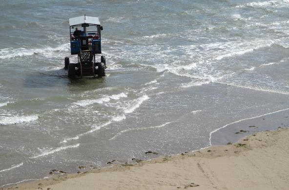 Auf den Strand fährt ein Fahrzeug zu, das momentan im ufernahen Wasser fährt und dicke Reifen hat und eine blaue offene Kabine. Es hat ein Gestell, das bei Flut hochgefahren werden kann, so dass es auch in höherem Wasser mobil bleibt.