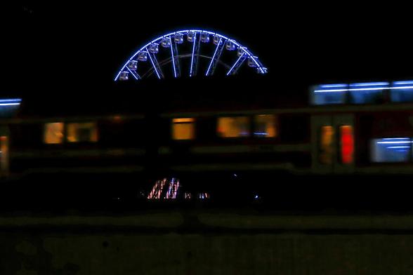 Abends, also finster.
Ein Riesenrad, beleuchtet.
Ein Zug fährt vorm Riesenrad vorbei.