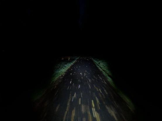 Dunkler Radweg mit verstreuten Blättern, umgeben von Gras, nur durch eine Lichtquelle beleuchtet