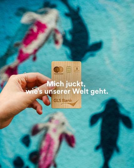 GLS Bank Holzkarte, die vor einem Wandgemälde mit Kois gehalten wird. Aufschrift: Mich juckt, wie's unserer Welt geht.