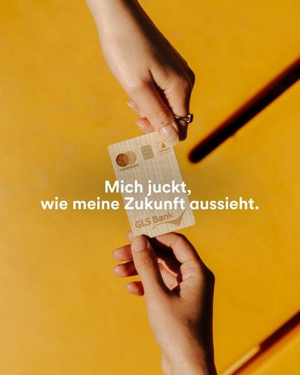 GLS Bank Holzkarte, die vor gelbem Hintergrund von zwei Händen gehalten wird. Aufschrift: Mich juckt, wie meine Zukunft aussieht.