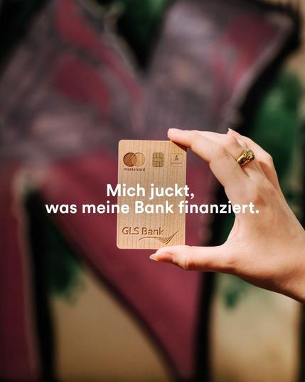 GLS Bank Holzkarte die vor einer Graffiti-Wand gehalten wird. Aufschrift: Mich juckt, was meine Bank finanziert.
