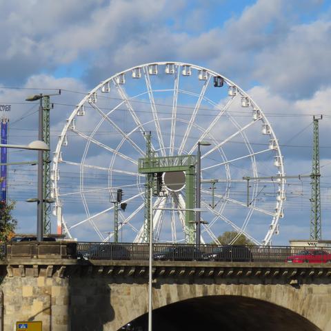 Weißes Riesenrad hinter alter Brücke.
Alle Gondeln, die sichtbar sind, sind weiß bis auf eine, die dunkelblau ist.