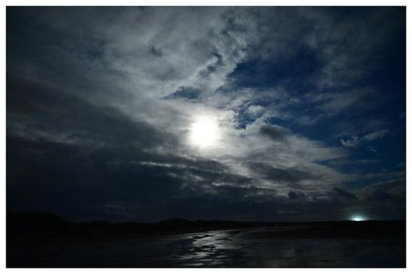 Das Bild zeigt eine nächtliche Landschaft mit einem teilweise bewölkten Himmel. Im Zentrum ist der Mond als ein heller, runder Lichtpunkt zu sehen. Die Wolken sind unterschiedlich dicht und bilden eine strukturierte Decke. Am unteren Bildrand spiegelt sich das Mondlicht auf einer Wasserfläche. Im Hintergrund sind dunkle, kaum erkennbare Dünen sichtbar.