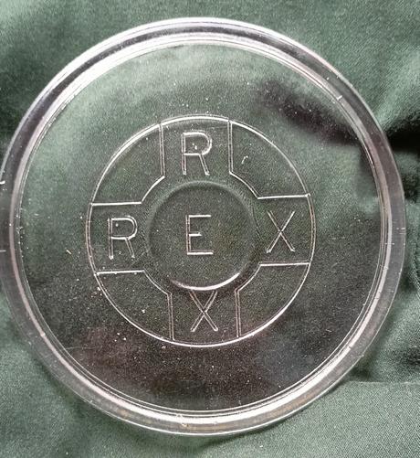 Glasdeckel mit den Buchstaben REX sowohl in horizontaler als in vertikaler Schreibung liegt auf einem grünen Kissen.