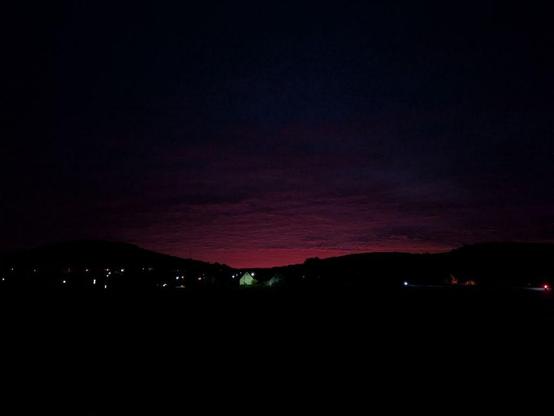 Dunkle Landschaft mit Hügeln und vereinzelten Lichtern unter einem rot-violetten Himmel bei Nacht