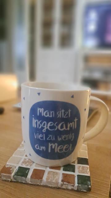Ein weiße Tasse  steht auf einem quadratischen Mosaik-Untersetzer auf einer Holzoberfläche. Auf der Seite des Bechers ist ein blauer Kreis mit dem Text „Man sitzt insgesamt viel zu wenig am Meer“ zu sehen. Um diesen blauen Kreis herum sind kleine blaue Dreiecke angeordnet. Der Untersetzer besteht aus kleinen quadratischen Fliesen in den Farben Braun, Weiß, Beige und Dunkelgrün. Der Hintergrund ist unscharf und zeigt Möbel sowie einen Bildschirm.