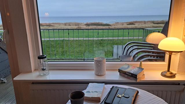 Ein Blick durch ein Fenster auf eine Küstenlandschaft unter einem bewölkten Himmel. Auf dem weißen Fenstersims stehen ein Glasbehälter, ein weißer Kerzenständer mit Muster, ein Buch und eine kleine, leuchtende Lampe. Hinter dem Fenster ist ein Metallgeländer zu sehen, das auf eine grüne Wiese und Dünen blickt, die zum Meer hin auslaufen. Im unteren Bildbereich befindet sich ein weißer Tisch mit einer dunklen Tasse, einem Buch und einer schwarzen Kladde.