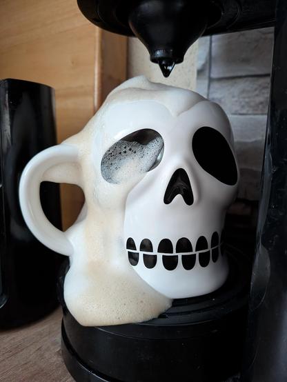 Eine Tasse in Form eines Totenschädels steht unter dem Auslass einer Kaffeemaschine. Weil zu viel Kaffee eingelaufen ist, läuft seitlich ordentlich weiß-bräunlicher Schaum außen an der Tasse herunter.
