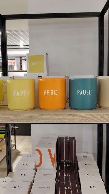 Mehrer bunte Tassen (gelb, orange, blau) in einem Regal. Auf ihnen steht "Happy", "Hero" oder "Pause". Weiter unten im Regal stehen weitere verpackte Becher.