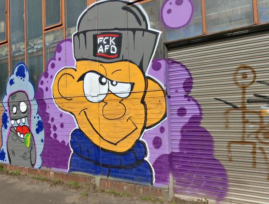 Graffiti an einer Wand zeigt eine große Comicfigur mit grauer Mütze, auf der 'FCK AFD' steht, und einem blauen Schal, daneben eine kleinere Comicfigur mit roter Zunge und grauer Mütze vor lila Hintergrund.