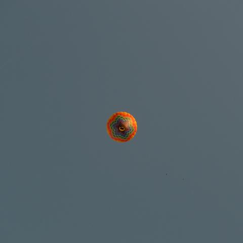 Inmitten einer quadratischen Farbaufnahme ist klein ein Heißluftballon zu sehen, der direkt von unten (90°) aufgenommen ist. Der Ballon ist orange und hat im unteren Bereich regenbogen-farbene Farbfelder. Der Korb befindet sich exakt in der Mitte der Aufnahme. Der wolkenlose Morgenhimmel ist graublau.