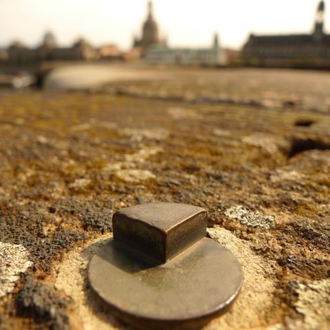 Makroaufnahme eines Metallstücks, aus dem ein Relief ragt, das wie ein Viertelstück eines Kuchens aussieht, gewölbt. Im Hintergrund unscharf die Frauenkirche, an einem heißen Sommertag.