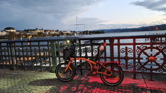 Mein bereits voll beleuchtetes, orangefarbenes Brompton Electric G-Line lehnt an einem geschmiedeten Geländer am Bürkliplatz mit Blick auf den Zürisee und — trotz Wolken — Alpenpanorama am Horizont.