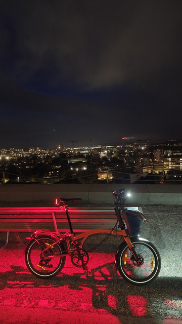 Mein voll beleuchtetes, orangefarbenes Brompton Electric G-Line lehnt an der Rückseite einer Bank am Nordhang des Züriberges mit nächtlichem Blick auf Schwamendingen, Oerlikon und am Horizont den hell beleuchteten Flughafen Zürich-Kloten.