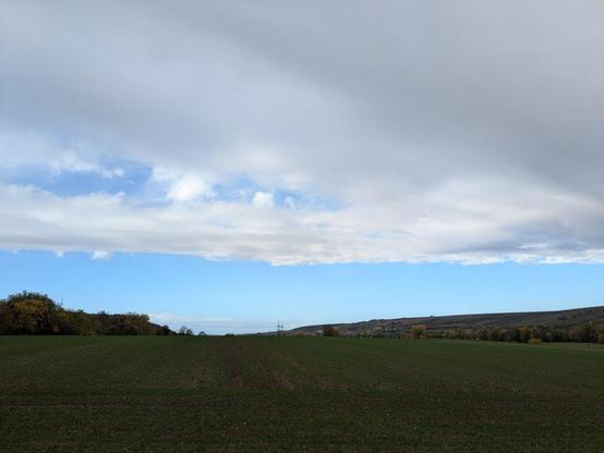Blick nach Norden beim Losfahren und damit Blick auf die klar abgegrenzte Wetterfront. Darunter weite Landschaft mit grünem Feld im Vordergrund.