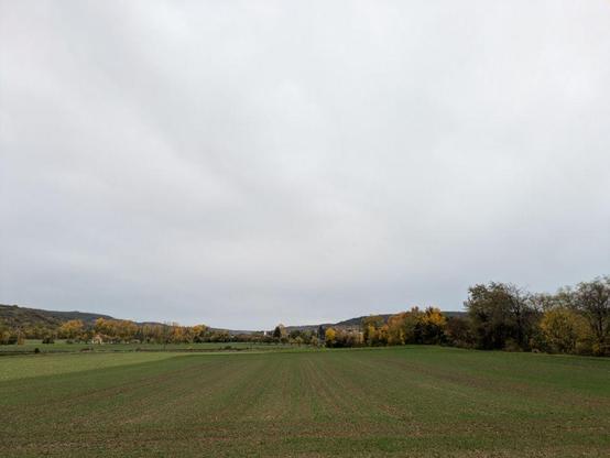 Blick nach Süden beim Losfahren auf weite Landschaft mit grünem Feld im Vordergrund und bewaldeten Hügeln im Hintergrund unter stark bewölktem Himmel