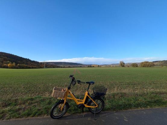 Nach einer Stunde Fahrt mein gelbes Fahrrad mit zwei Körben steht auf Asphaltweg vor weitem grünen Feld und blauem Himmel