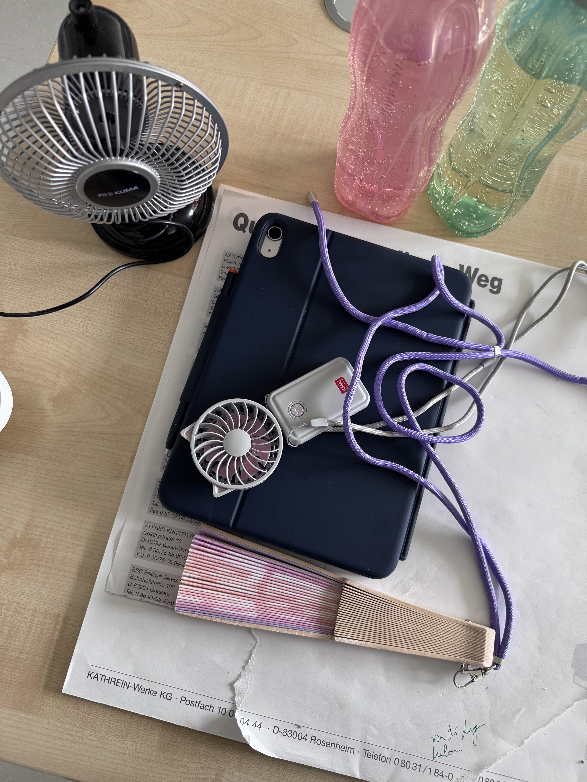 Auf dem Schreibtisch sind zu sehen:
Ein USB-Ventilator, ein Legami Ventilator zum umhängen, ein bunter Fächer mit Schnur zum umhängen und zwei Wasserflaschen